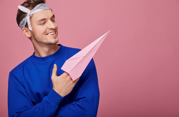Sorrindo cara feliz no suéter azul escuro bandanin branco com avião de papel rosa na mão