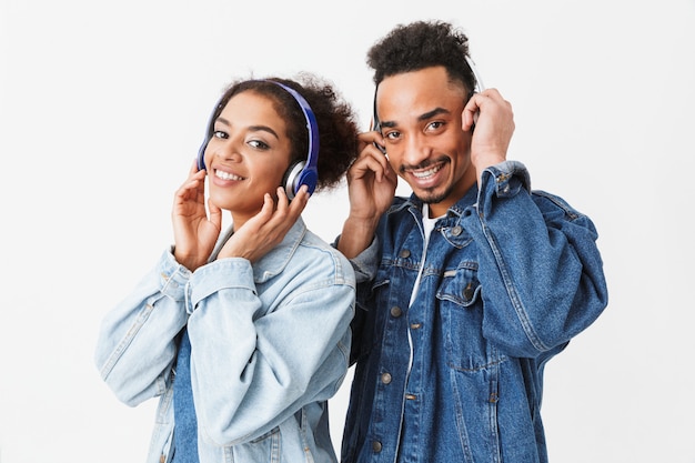 Sorrindo casal Africano em camisas jeans, ouvir música por fones de ouvido ao longo da parede cinza