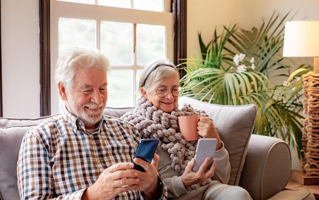 Sorrindo casal sênior relaxando sentado no sofá juntos na sala de estar usando smartphones