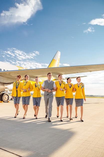 Sorrindo comissários de bordo de homens e mulheres vestindo uniforme de aviação enquanto caminhava perto de avião de passageiros no aeroporto