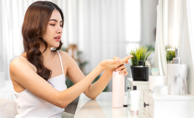 Sorrindo de uma jovem linda mulher asiática limpa pele branca saudável fresca olhando para o espelho. menina asiática segurando loção corporal e aplicando creme hidratante em casa