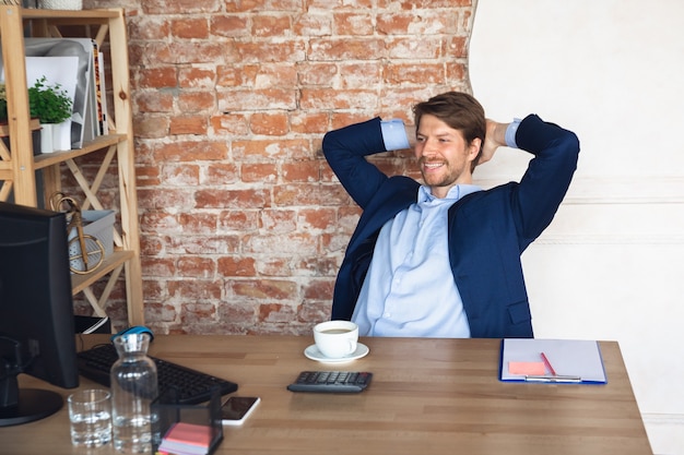 Sorrindo, descansando, parece encantado, bem sucedido. Jovem, gerente, volta ao trabalho em seu escritório após a quarentena, sente-se feliz inspirado. Voltando à vida normal. Negócios, finanças, conceito de emoções.