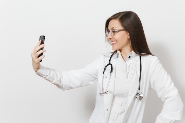 Sorrindo, diversão linda jovem médica com estetoscópio, óculos isolados no fundo branco. médica em vestido de médico fazendo selfie no celular. pessoal de saúde, saúde, medicina concentrada