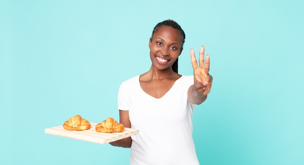 Sorrindo e parecendo amigável, mostrando o número três e segurando uma bandeja de croissant