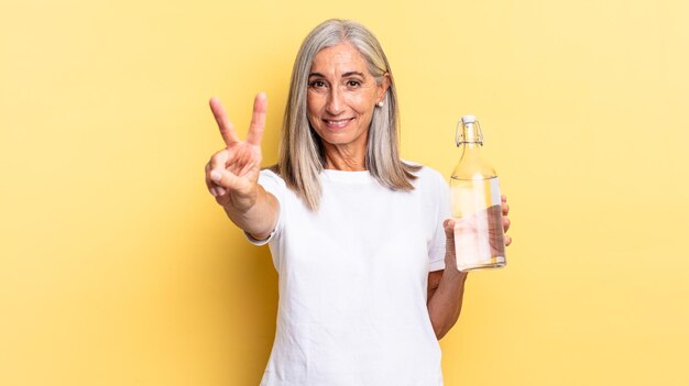 Sorrindo e parecendo feliz, despreocupado e positivo, gesticulando vitória ou paz com uma mão e segurando uma garrafa de água