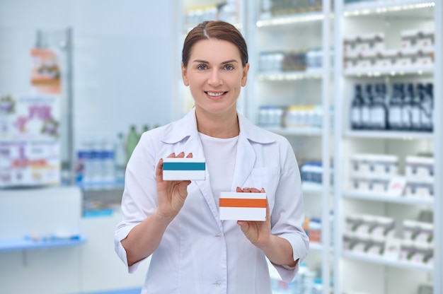 Sorrindo e satisfeita, linda farmacêutica feminina segurando duas caixas de papelão com remédios na frente dela