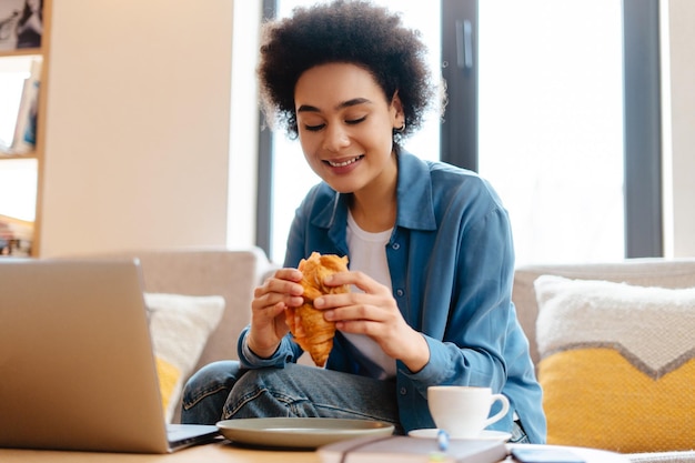 Sorrindo elegante mulher afro-americana com cabelo encaracolado freelancer segurando croissant fresco