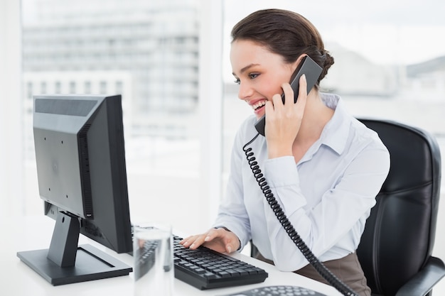 Sorrindo empresária usando telefone fixo e computador no escritório