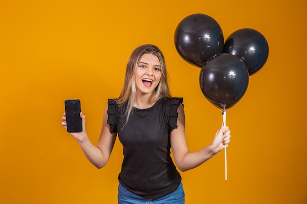 Sorrindo encantadora jovem segurando um celular com tela em branco vazia sobre fundo amarelo com retrato de estúdio de balões de ar. liquidação da black friday