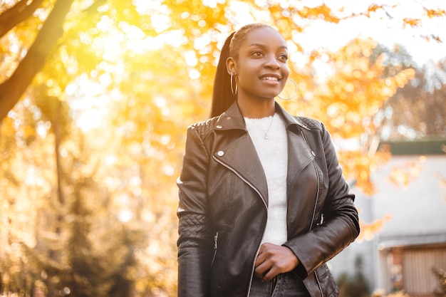 Sorrindo espantada mulher afro-americana vestida de jaqueta de couro preta em pé no parque da cidade