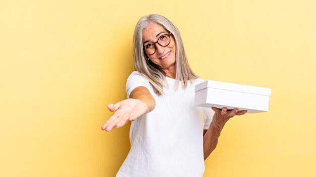 Sorrindo feliz com olhar amigável, confiante e positivo, oferecendo e mostrando um objeto ou conceito e segurando uma caixa branca