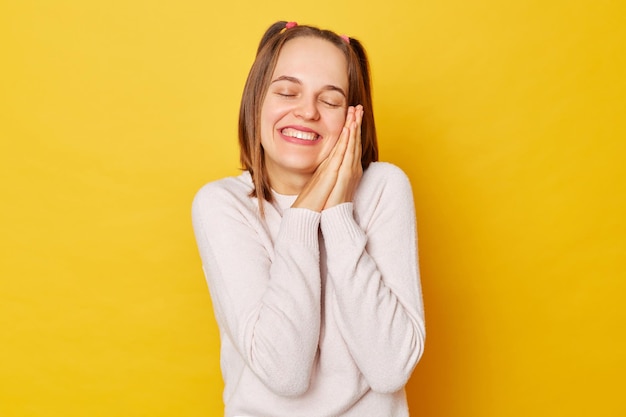 Foto sorrindo, feliz e relaxante adolescente em jumper com rabos de cavalo isolado sobre fundo amarelo, aproveitando a soneca de fim de semana, apoiando-se nas mãos, mantém os olhos fechados