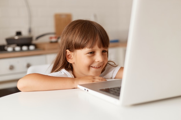 Sorrindo feliz garotinha pré-escolar com cabelo escuro, sentada na frente do computador laptop na cozinha, olhando para o display, expressando emoções positivas, assistindo desenhos animados.