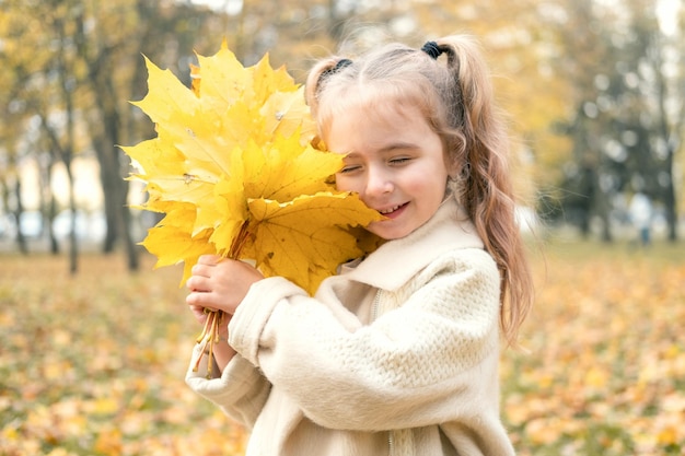 sorrindo feliz menina de casaco e vestido segurando folhas de outono, se divertindo na floresta de outono