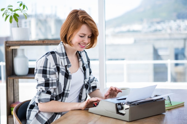 Sorrindo hipster mulher sentada em uma mesa, digitando na sua máquina de escrever