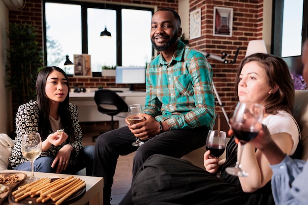 Sorrindo homem afro-americano sentado no sofá enquanto celebra a promoção do emprego com os melhores amigos. Grupo diversificado de pessoas fazendo festa de vinho em casa para comemorar o aniversário da amizade.
