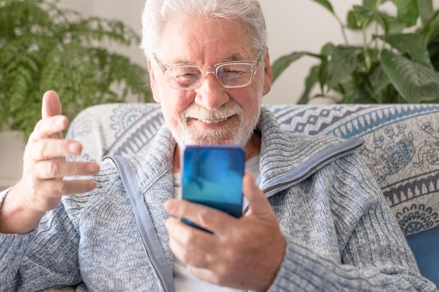 Sorrindo homem barbudo idoso sentado no sofá em casa enquanto fala no celular gesticulando com a mão Sênior em óculos e suéter azul claro