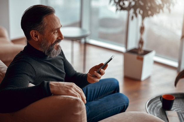 Sorrindo homem barbudo sentado na poltrona e lendo mensagem no smartphone