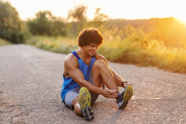 Sorrindo homem musculoso com cabelos cacheados, sentado na estrada e amarrar o cadarço. Dia ensolarado de verão.