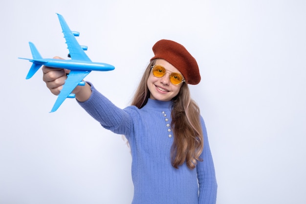 Sorrindo, inclinando a cabeça, linda garotinha de óculos com chapéu segurando um avião de brinquedo