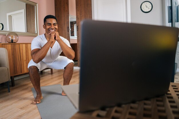 Sorrindo jovem afro-americano vestindo camiseta branca exercitando e fazendo agachamentos online na frente do laptop na sala doméstica brilhante. conceito de treinamento esportivo no ginásio em casa.