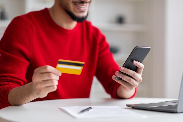 Sorrindo jovem árabe com barba mostrando cartão de crédito e digitando no smartphone no escritório em casa