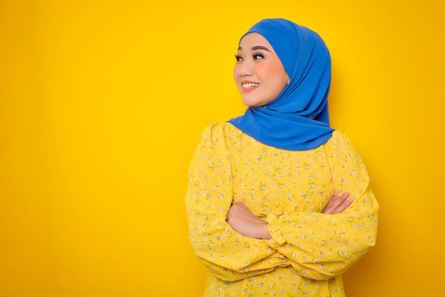 Sorrindo jovem asiática em vestido casual de pé com braços cruzados olhando para longe isolado em fundo amarelo