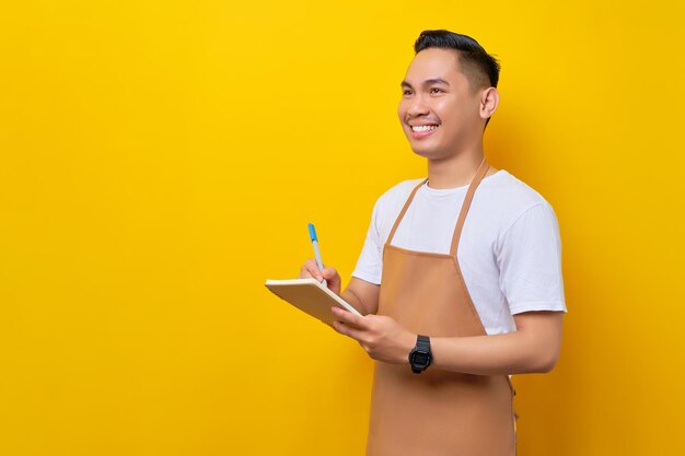 Sorrindo jovem asiático barista barman empregado vestindo avental marrom trabalhando na cafeteria anote o menu do pedido no caderno olhe de lado isolado no fundo amarelo Conceito de inicialização de pequenas empresas