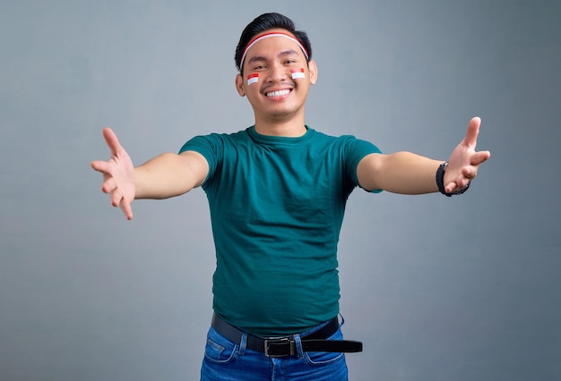 Sorrindo jovem asiático de pé com as mãos estendidas para abraços isolados no conceito de celebração do dia da independência da indonésia de fundo cinza