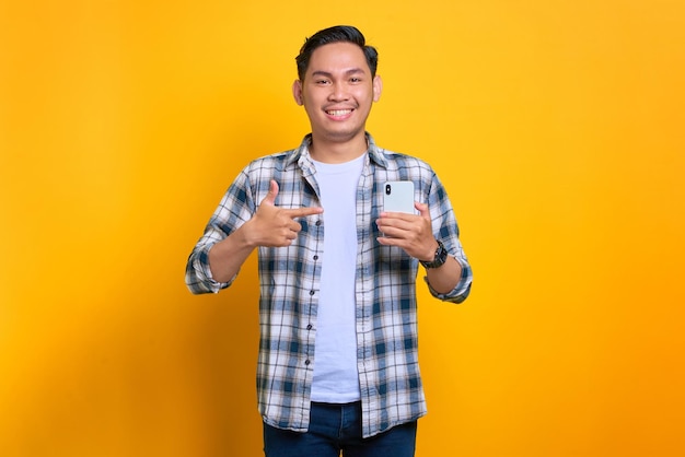 Sorrindo jovem asiático em camisa xadrez apontando dedos para um telefone celular recebendo boas notícias isoladas em fundo amarelo