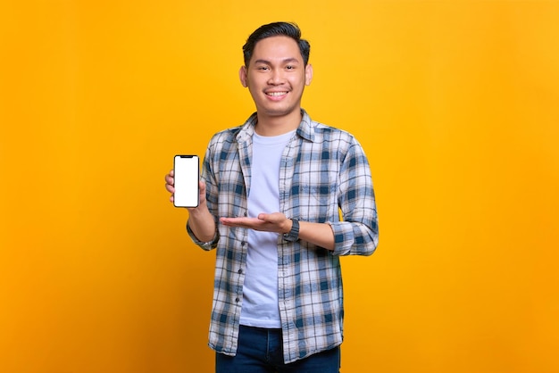 Sorrindo jovem asiático em camisa xadrez mostrando a tela em branco do celular recomendando aplicativo isolado em fundo amarelo