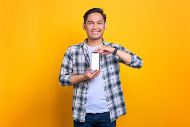 Sorrindo jovem asiático em camisa xadrez mostrando a tela em branco do celular recomendando aplicativo isolado em fundo amarelo