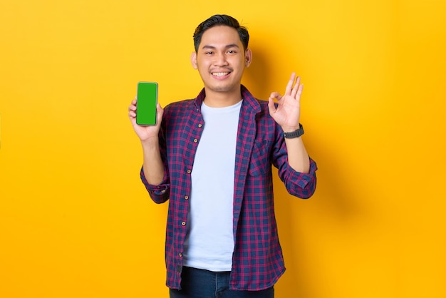 Sorrindo jovem asiático em camisa xadrez mostrando smartphone de tela em branco e gesticulando sinal de ok isolado em fundo amarelo