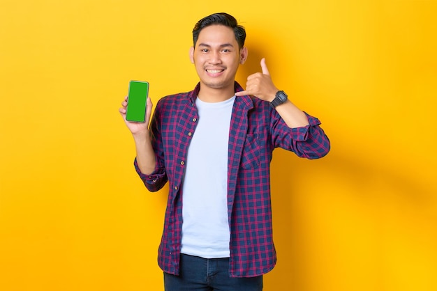 Sorrindo jovem asiático em camisa xadrez mostrando smartphone de tela em branco e sinal de telefone gesticulando isolado em fundo amarelo