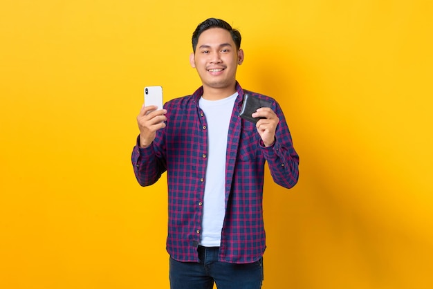 Sorrindo jovem asiático em camisa xadrez segurando telefone celular e mostrando carteira isolada em fundo amarelo