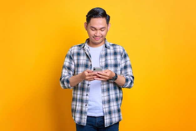 Sorrindo jovem asiático em camisa xadrez usando telefone celular lendo boas notícias isoladas em fundo amarelo
