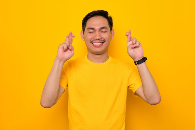 Sorrindo jovem asiático em camiseta casual cruzando os dedos com os olhos fechados esperando por sorte isolada em fundo amarelo Conceito de estilo de vida de pessoas