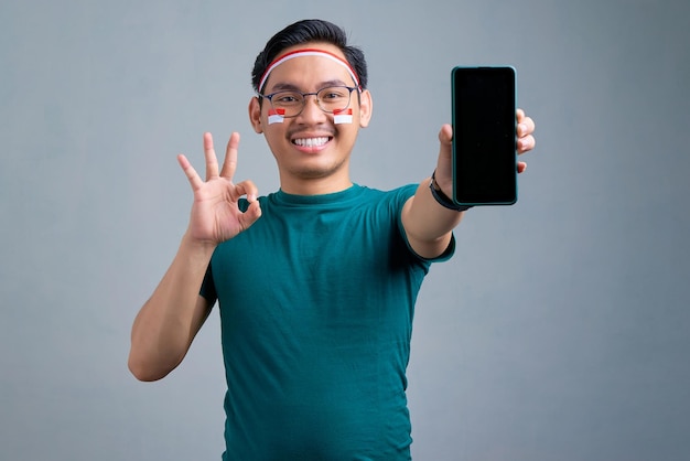 Sorrindo jovem asiático em camiseta casual mostrando celular de tela em branco e gesticulando sinal de ok isolado no conceito de celebração do dia da independência indonésia de fundo cinza