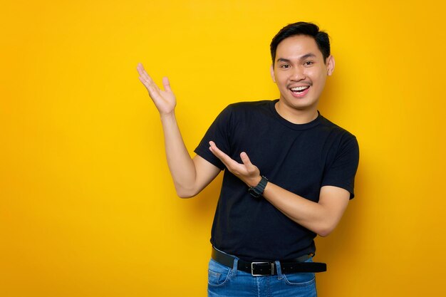 Sorrindo jovem asiático em camiseta casual segurando espaço de cópia na palma da mão para inserir anúncio isolado em fundo amarelo Conceito de estilo de vida de pessoas