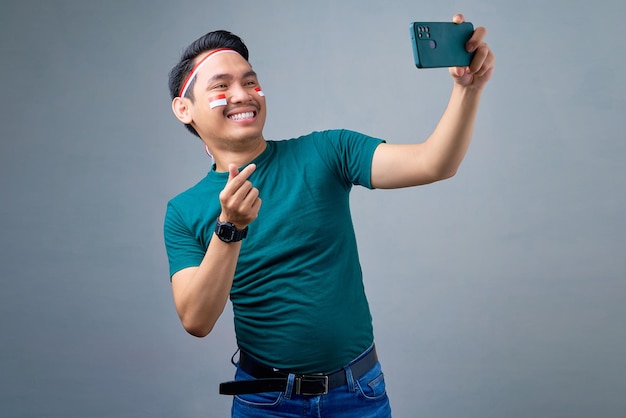 Sorrindo jovem asiático tomando selfie no celular mostrando coração coreano com dois dedos cruzados isolados em fundo cinza conceito de celebração do dia da independência indonésia