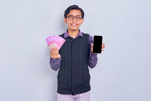 Sorrindo jovem asiático vestindo camisa mostrando a tela em branco do celular e segurando a nota de dinheiro em dinheiro rupia isolada no fundo branco