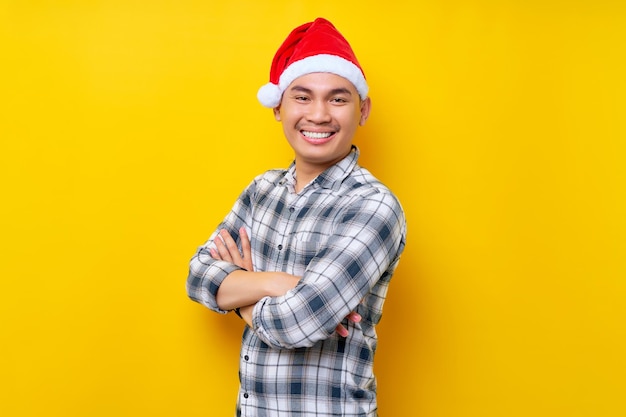 Sorrindo jovem asiático vestindo uma camisa xadrez em um chapéu de Natal em pé de mãos dadas cruzadas e dobradas parece câmera na celebração de fundo amarelo feriado de Natal e conceito de ano novo