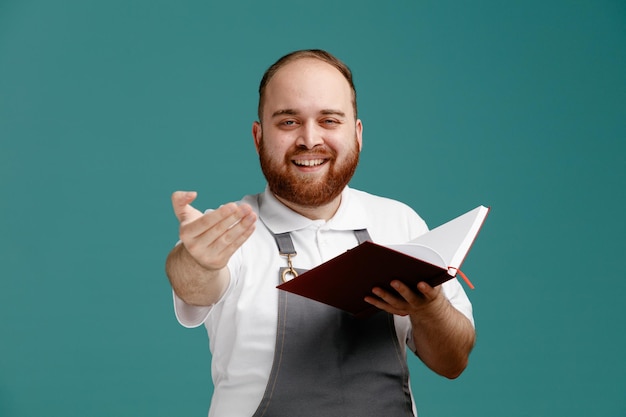 Sorrindo jovem barbeiro masculino vestindo camisa branca e avental de barbeiro segurando o bloco de notas olhando para a câmera mostrando venha aqui gesto isolado no fundo azul