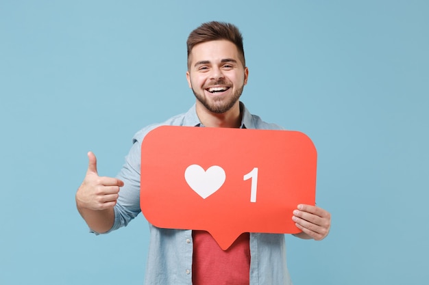 Foto sorrindo jovem barbudo em camisa casual posando isolado em fundo azul pastel. conceito de estilo de vida de pessoas. mock up espaço de cópia. segure enorme como o sinal do formulário do coração da rede social, mostrando o polegar para cima.