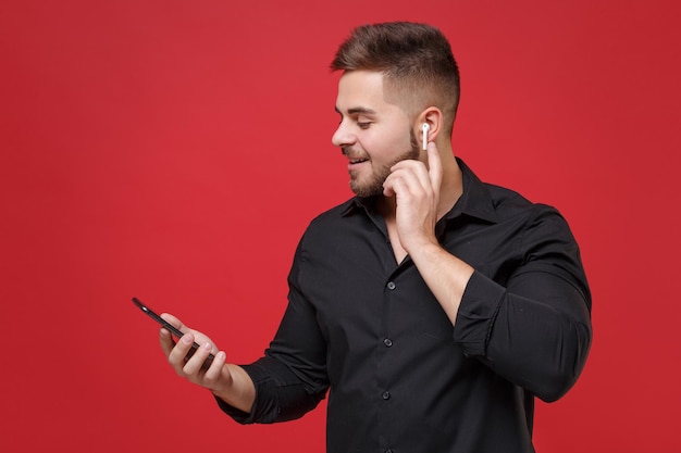 Sorrindo jovem barbudo na camisa preta clássica posando isolado no retrato de estúdio de fundo de parede vermelha brilhante. Conceito de estilo de vida de emoções de pessoas. Mock up espaço de cópia. Usando cápsulas de ar e telefone celular.