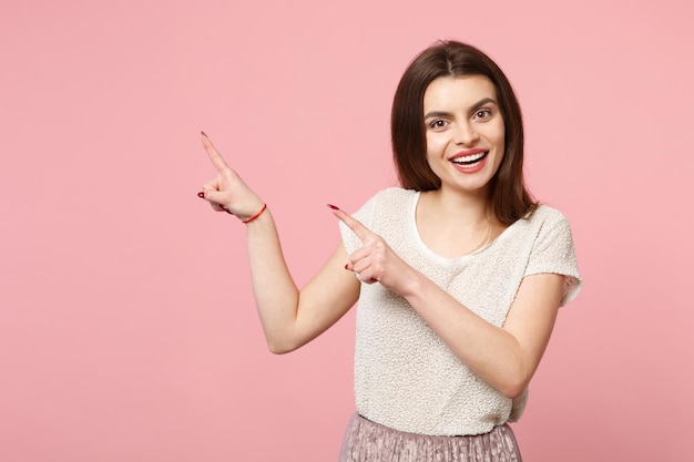 Sorrindo jovem bonita em roupas leves casuais posando isolado no fundo da parede rosa, retrato de estúdio. Conceito de estilo de vida de emoções sinceras de pessoas. Mock up espaço de cópia. Apontando os dedos indicadores para o lado.