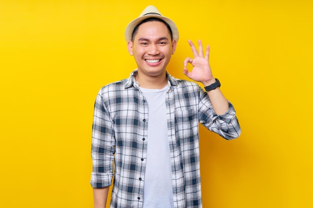 Sorrindo jovem bonito étnico asiático de 20 anos vestindo roupas casuais chapéu confiante e mostrando ok ok gestos isolados no fundo amarelo Conceito de estilo de vida de pessoas