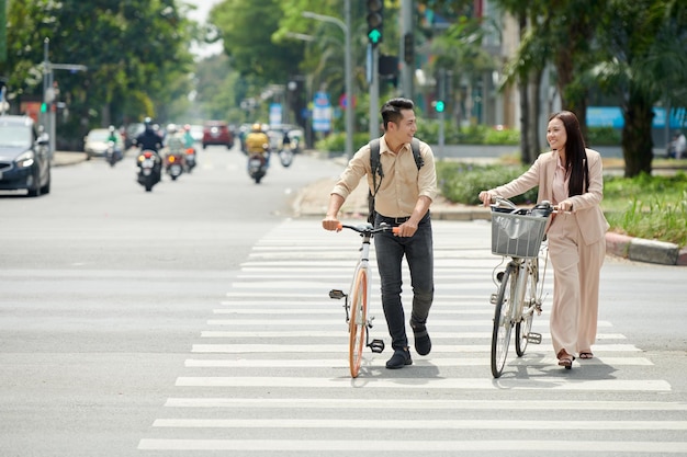 Sorrindo jovem casal asiático com bicicletas atravessando a estrada na faixa de pedestres