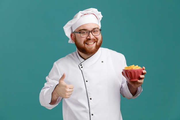 Sorrindo jovem chef masculino vestindo óculos uniforme e boné mostrando a tigela de macarrão olhando para a câmera mostrando o polegar isolado no fundo azul