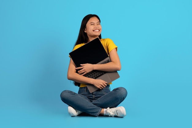 Sorrindo jovem chinesa em casual sentado no chão e abraçando laptop com tela em branco isolada